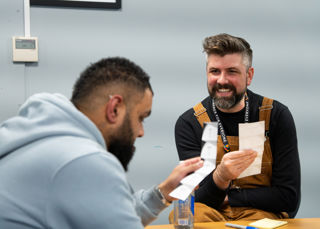 One person is reading off a small piece of paper, another person looks at them. They are both smiling.